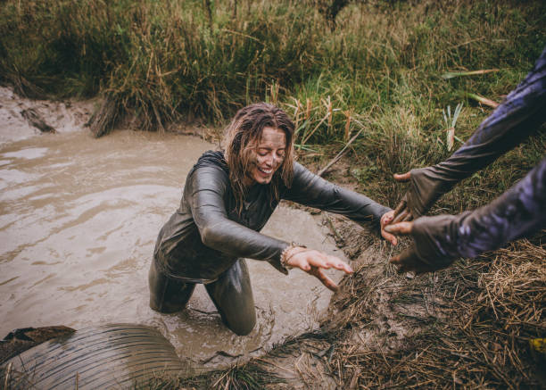 Help Me Out! Woman is being pulled out of a ditch full of muddy water by her friend. They are both taking part in a charity obstacle course. obstacle course stock pictures, royalty-free photos & images