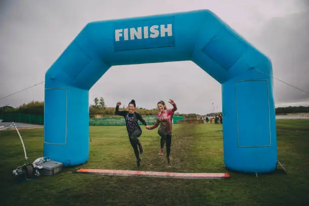 Two women are holding hands and skipping over the finish line of a charity obstacle course.