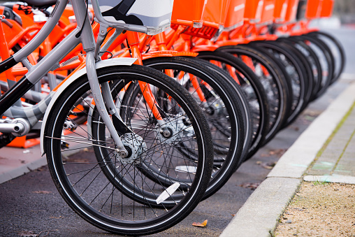 Orange bikes with baskets in front and polished wheels for paid public use stay lined and one gray bike symbolic not likely other stand out from the rest of this series.
