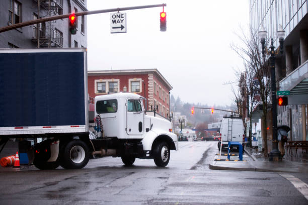 Day cab semi truck with dark blue dry van semi trailer turn on urban city street delivering food and beverages. Modern compact powerful semi truck with a semi trailer frame fitted by a dark rubberized textile, turn at the crossroads with traffic lights in modern Portland downtown, delivering food and beverages to stores and restaurants. truck in rain stock pictures, royalty-free photos & images