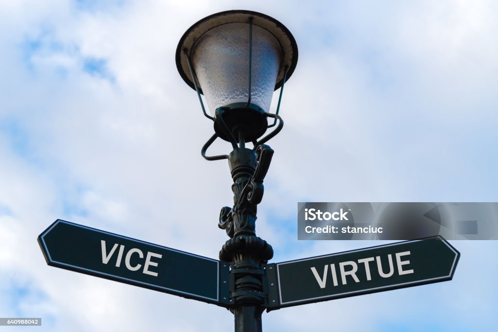 Vice versus Virtue directional signs on guidepost Street lighting pole with two opposite directional arrows over blue cloudy background. Vice versus Virtue concept. Arrow Symbol Stock Photo