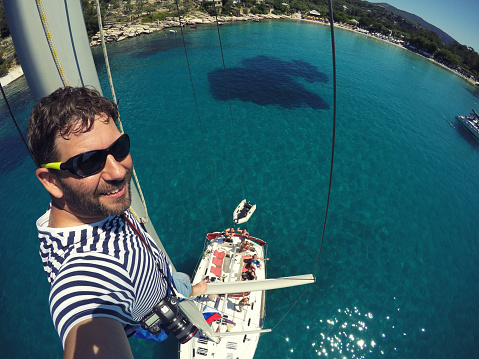 One happy man making selfie from the top of the sailboat mast above the crystal clear water. He is wearing t-shirt with blue and white stripes, which is traditional sailor pattern. Some of the people from crew are in the water, enjoying at sunny day. Photo taken with GoPro camera