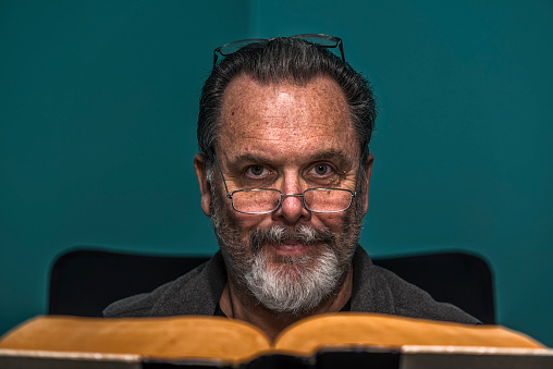 A portrait of a middle aged Caucasian male two pair of wearing reading eyeglasses looking at the camera while reading a large vintage book similar to an atlas. He has an unkempt graying beard, with his hair slicked back.