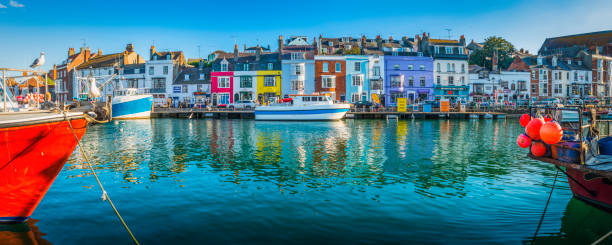 Fishing boats moored in busy harbour colourful cottages Weymouth Dorset Colourful cottages, fishing boats and yachts around the harbour as holiday makers and locals enjoy the warm summer evening outside waterfront pubs and restaurants at the popular seaside resort town of Weymouth, Dorset. dorset england photos stock pictures, royalty-free photos & images