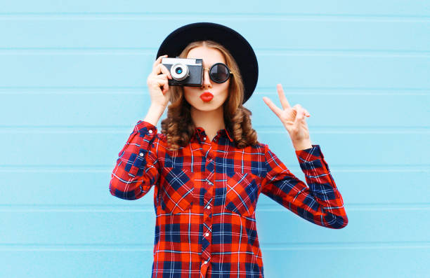 fashion pretty young woman blowing red lips with retro camera wearing a black hat, red checkered shirt over blue background - photographer enjoyment elegance old fashioned imagens e fotografias de stock