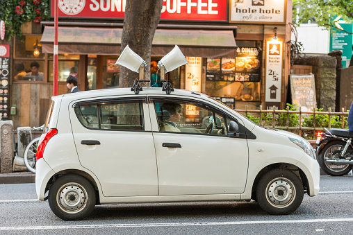 In the Shibuya Ward of Tokyo, Japan a man drives an unmarked white four door hatchback car with two audio loud speakers attached to the roof. The speakers broadcast recorded advertisements in Japanese.