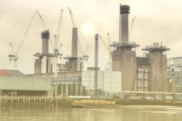 An image of Battersea Power Station displaying building cranes and construction work in and around the property.  Thames River in foreground. Sun in Background.