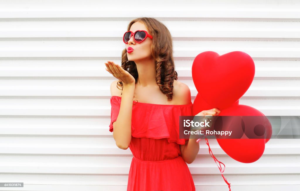 Portrait woman in red dress sends air kiss with balloon heart shape over white background Valentine's Day - Holiday Stock Photo