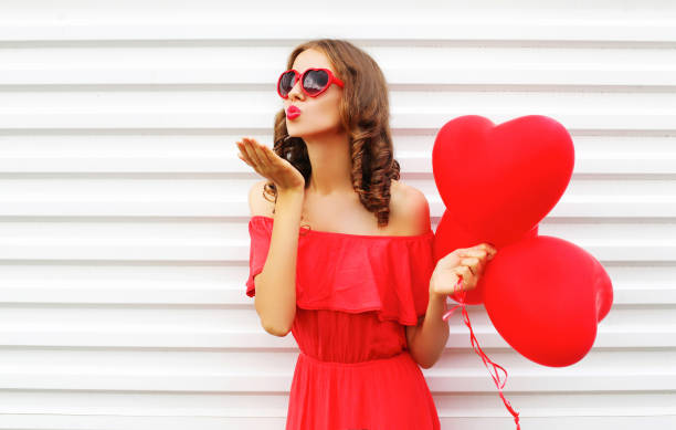 Portrait woman in red dress sends air kiss with balloon heart shape over white background stock photo