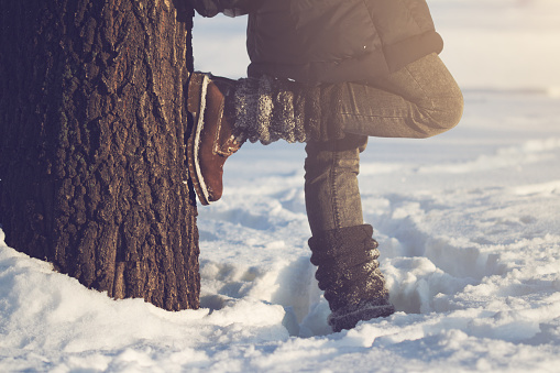 horizontal close up of woman wearing brown boots, textile gaiters and blue jeans leaning against a large tree covered in snow in winter time, concept for hiking