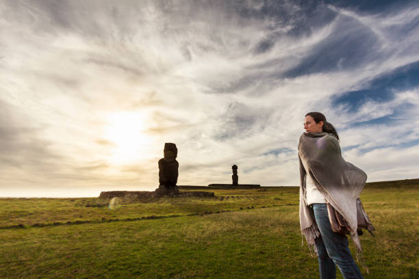 Woman Traveling in Easter Island A DSLR Canon photo of a Brazilian caucasian beautiful woman traveling in Easter Island, Chile. She is standing at a huge open green field with two Moai statues in background. The sun is low in a dramatic sky behind white clouds. chile tourist stock pictures, royalty-free photos & images