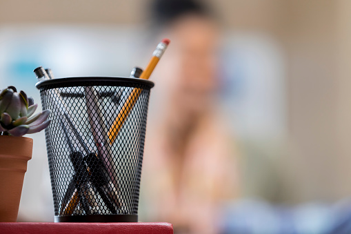 Close up of pencil cup on a desk in a modern office. A succulent plant is also on the desk. A woman is blurred in the background. Copy space is available.