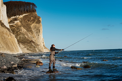 Canidid photo of a woman, waist deep in water who casting out her fishing line next to the cliffs at Møns Klint on the island of Møn in Denmark. Girls fish too is her motto! Colour, horizontal format with lots of copy space.