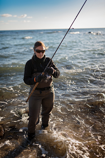 Canidid photo of a woman, walking back to the shore holding her fishing rod, after fishing in waist deep in water  below the cliffs at Møns Klint on the island of Møn in Denmark. Girls fish too is her motto! Colour, portrait format with some copy space.