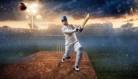 Cricket batsman bouncing a ball. He is wearing unbranded sports cloth and equipment. The bleachers full of people are blurred behind the player. There is a rain falling from the dark skies and intentional lenseflares on the image. The stadium is made in 3D.