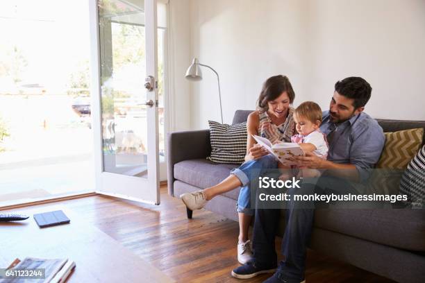 Parents Reading With Baby Daughter On Sofa At Home Stock Photo - Download Image Now - Family, Indigenous Peoples of the Americas, Domestic Life