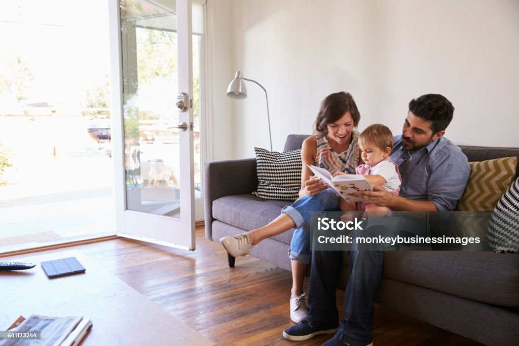 Pais lendo em casa com a filha bebê no sofá - Foto de stock de Família royalty-free