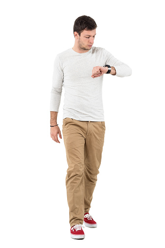 Stylish young man walking and checking time on wrist watch. Full body length portrait isolated over white studio background.