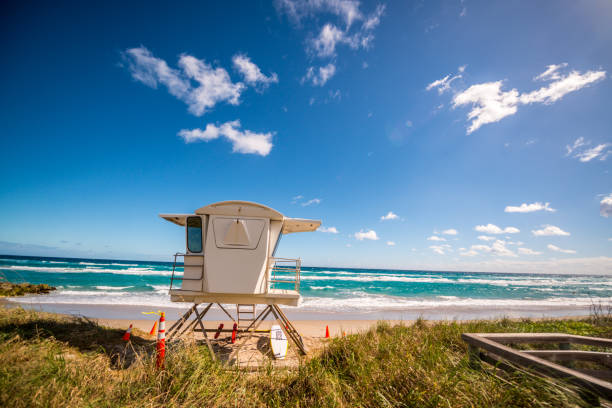 Empty white lifeguard chair on an empty beach with  orange life buoys hanging on the side.The chair stands against a clear blue sky in the sand.