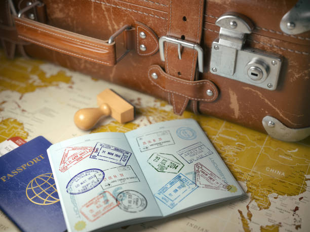 An open passport with colorful visa stamps rests on a world map beside a blue passport, a rubber stamp, and a vintage leather suitcase.