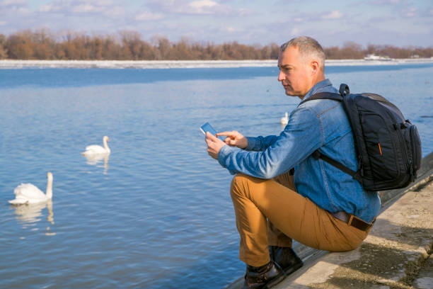 lonely man sitting by river with backpack and digital tablet - travel ipad isolated backpack imagens e fotografias de stock