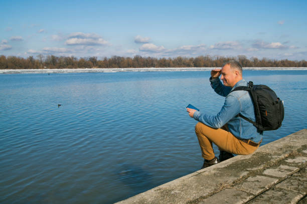 homem maduro sentado junto a um lago admirar a vista - travel ipad isolated backpack imagens e fotografias de stock