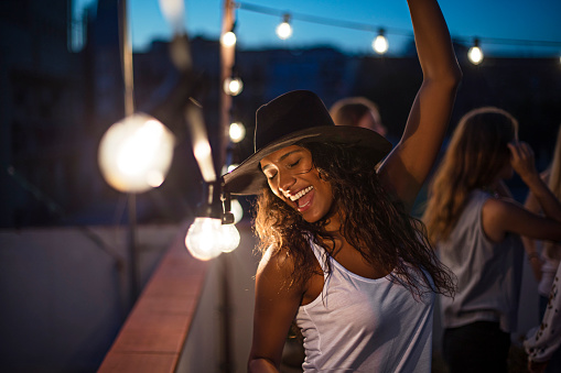 Cheerful woman dancing with arm raised during party. Happy female is enjoying on illuminated terrace. Friends are celebrating during dusk.