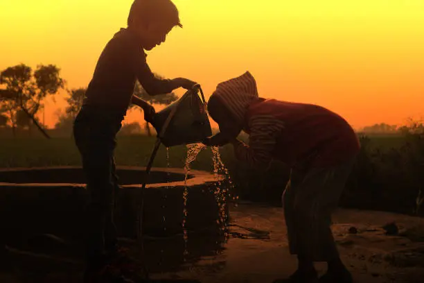 Village children drinking water at water well in the dusky day during sunset.