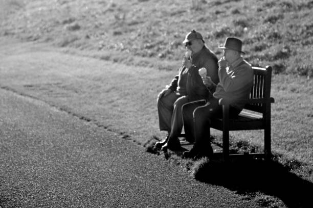 Two senior men in hats eating ice cream cones on a seat beside Seaton Sluice beach with grass bank in the background.