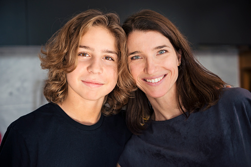 Cute teenage son with long curly hair is posing againts his beautiful mother at home during a family reunion. Their heads touch and both are smiling and looking at the camera. Horizontal head and shoulders indoors shot with copyspace. This was taken in Quebec, Canada.