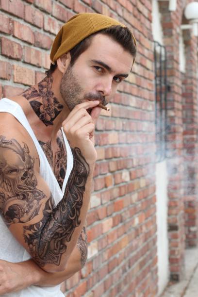 uomo barbuto tatuato che fuma una sigaretta all'aperto - stubble men tattoo sensuality foto e immagini stock