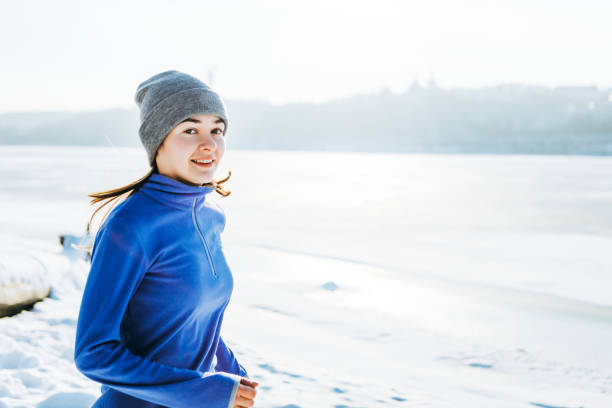 Young sporty woman running along the riverbank at a frozen river on a sunny winter day wearing tights and fleece jacket