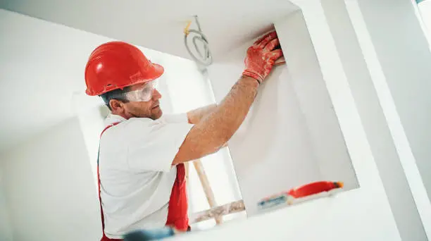 Closeup side view of a construction worker sanding a drywall and preparing it for a paint job. He's paying special attention to the corners and angles.The man is wearing red protective uniform and a red hardhat.