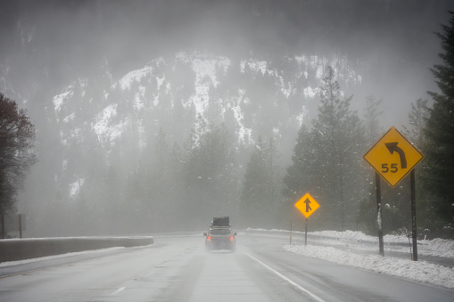 A heavily loaded up family car is driving along a freeway. A forest lines both sides of the road, and a mountain looms in the background. It's icy cold weather, with it snowing. The setting is located on the border between Northern California and Southern Oregon in the Pacific Northwest during Winter.