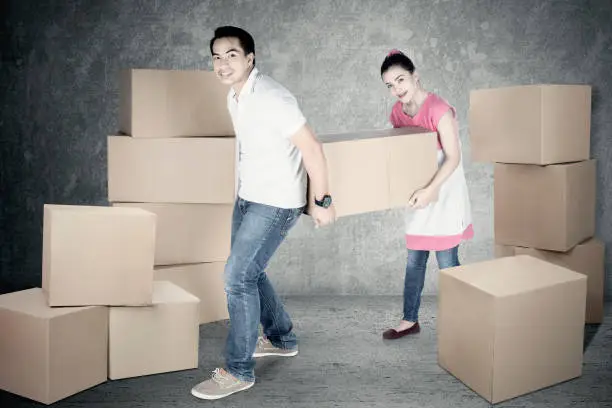 Full length of young husband and wife moving box together while looking at the camera