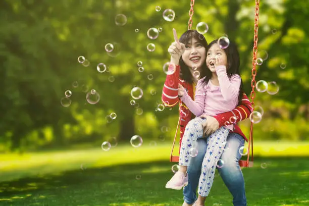 Portrait of young mother and cute child sitting on the swing while pointing at soap bubbles in the air