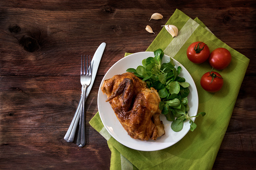 grilled half chicken with tomatoes, corn salad and garlic, white plate, green napkin and silverware on a dark rustic wooden table with copy space