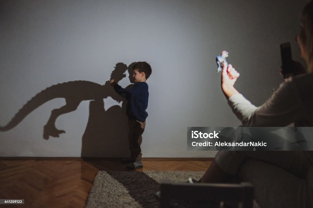 Little dinosaur lover Little boy and his mother are playing with shadow puppets on the wall by using dinosaur toys Shadow Stock Photo