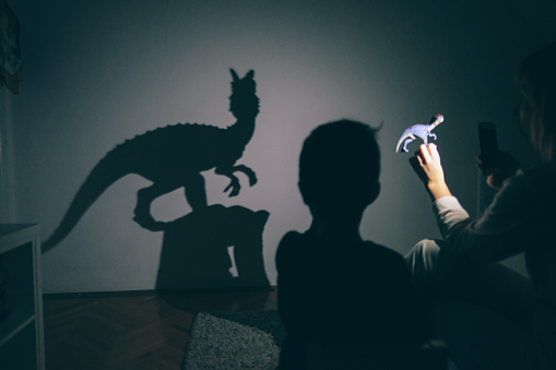 Little boy and his mother are playing with shadow puppets on the wall by using dinosaur toys