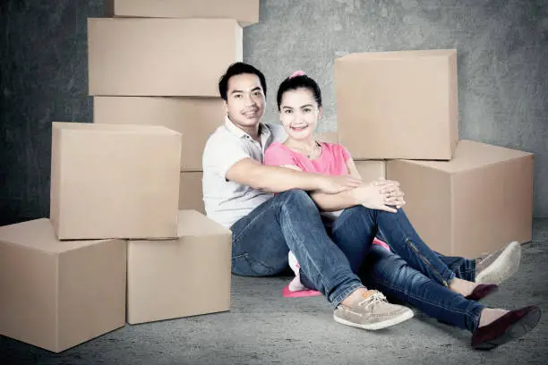 Portrait of young husband and wife sitting on the floor with stack of boxes while looking at the camera