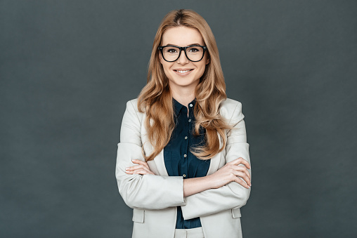 Gorgeous woman in smart casual wear keeping arms crossed and looking at camera while standing against grey background