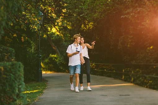 Teen couple walking in the park and talking. Female pointing and male holding smart phone.