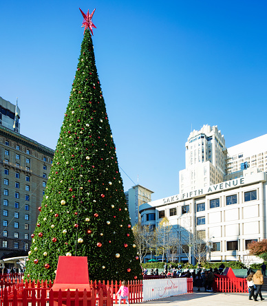 San Francisco Macy's Christmas tree in Union Square on a sunny day, A little girl dances in front of it while several people are visiting the site in its shadow. Saks Fifth Avenue store in the background.