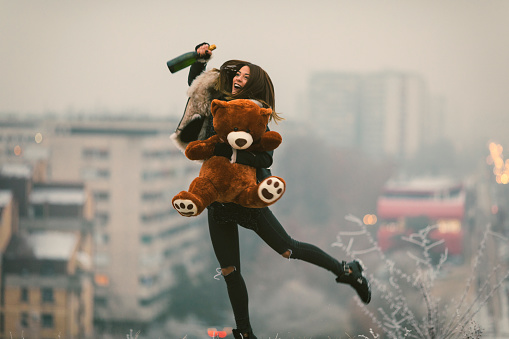 Happy Young Woman Having Fun Outdoors on the top of the hill. Hodling  champagne and teddy bear with the city in fog in background