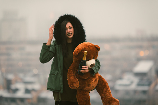Happy Young Woman Having Fun Outdoors on the top of the hill. Hodling  champagne and teddy bear with the city in fog in background