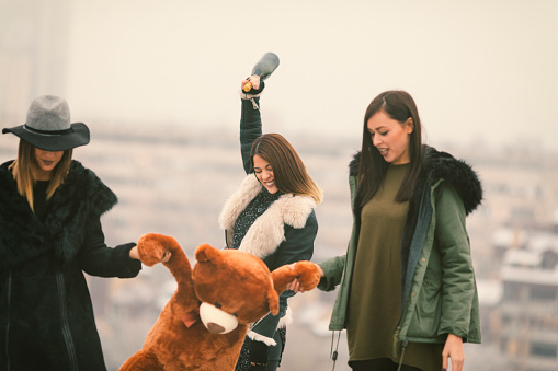 Happy Young Women Having Fun Outdoors on the top of the hill. Hodling  champagne and teddy bear with the city in fog in background