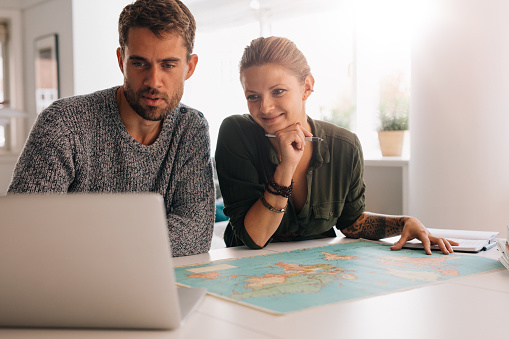 Young man and woman looking at laptop computer with world map in front. Couple using technology to explore the world.