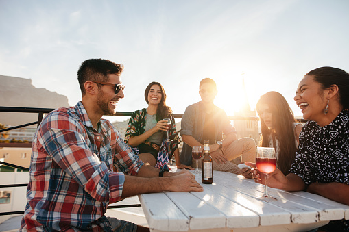 Multiracial group of friends having cocktail party on the rooftop. Young men and women sitting around table with drinks and laughing.