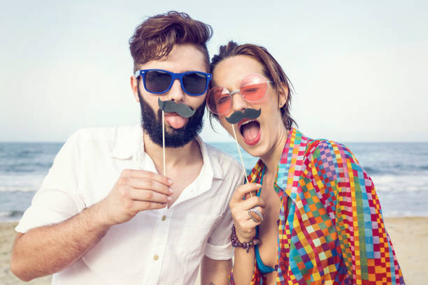 Fake mustache fun Couple on the beach posing with paper mustaches and colorful glasses women movember mustache facial hair stock pictures, royalty-free photos & images