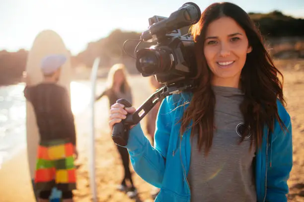 Young smiling woman holding video camera, having a photo shoot with her friends at the beach, while they are preparing to surf